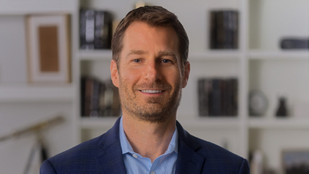 Fynanc Bio Pics_Jordan Cooper Jordan Cooper, a Fynanc Coach, investor, and financial strategist, smiling confidently in a professional setting with bookshelves in the background.