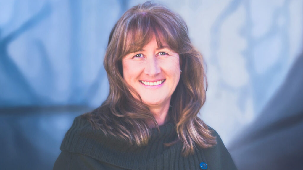 Fynanc Bio Pics_Sonja Reichenberg Sonja Reichenberg, a Fynanc Coach and Performance and Accountability Coach, smiling warmly in a professional setting with a soft, blue-toned background.